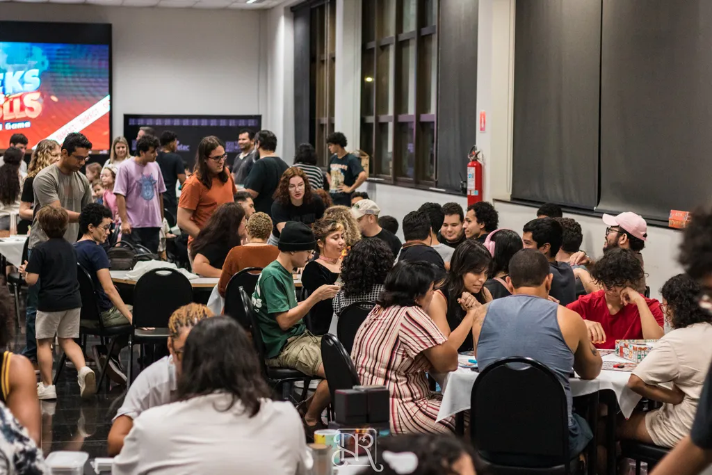 Salão fechado e cheio de pessoas sentadas ao redor de mesas cobertas com toalhas brancas, concentradas em jogos de tabuleiro e de cartas. O público é variado, com adultos, jovens e crianças. Ao fundo, mais pessoas conversam em pé e há um telão colorido iluminando o ambiente.